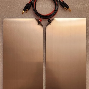 May include: Two rectangular, brushed metal plates with a smooth, reflective surface. Each plate features a small, angled corner tab with a single electrical terminal, one red and one black. A coiled black and red electrical cable with gold-colored banana plug connectors is positioned above the plates.