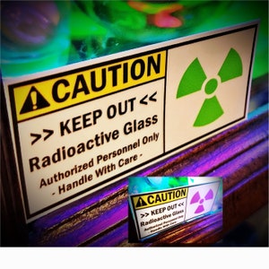 May include: A yellow and black caution sign with a green radiation symbol. The sign reads "CAUTION >> KEEP OUT << Radioactive Glass Authorized Personnel Only - Handle With Care -"