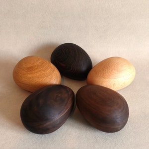 May include: Five wooden eggs in a variety of wood types and colours. Two are dark brown, two are light brown, and one is a dark brown with a wood grain pattern.