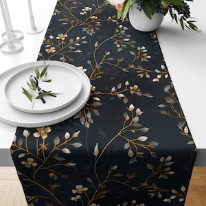 Puede incluir: Un camino de mesa negro con un patrón floral dorado y blanco. El camino de mesa presenta ramas delicadas con pequeñas flores blancas y hojas doradas.