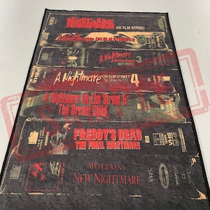 May include: A black and red rug with a design of VHS tapes featuring the Nightmare on Elm Street movie franchise. The rug features the titles of the movies, including "A Nightmare on Elm Street", "A Nightmare on Elm Street 2", "A Nightmare on Elm Street 3: Dream Warriors", "A Nightmare on Elm Street 4: The Dream Master", "A Nightmare on Elm Street 5: The Dream Child", "Freddy's Dead: The Final Nightmare", and "Wes Craven's New Nightmare".