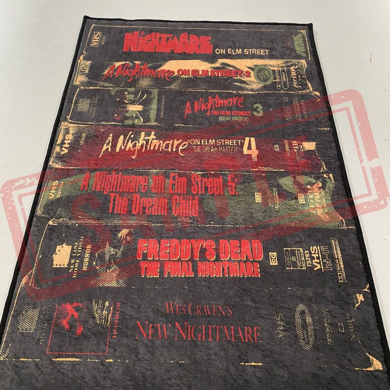 May include: A black and red rug with a design of VHS tapes featuring the Nightmare on Elm Street movie franchise. The rug features the titles of the movies, including "A Nightmare on Elm Street", "A Nightmare on Elm Street 2", "A Nightmare on Elm Street 3: Dream Warriors", "A Nightmare on Elm Street 4: The Dream Master", "A Nightmare on Elm Street 5: The Dream Child", "Freddy's Dead: The Final Nightmare", and "Wes Craven's New Nightmare".