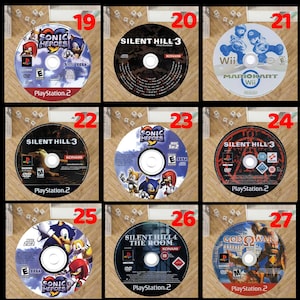Pode incluir: Uma cole&ccedil;&atilde;o de discos de videojogos, incluindo t&iacute;tulos como "Sonic Heroes", "Silent Hill 3" e "Mario Kart Wii". Os discos s&atilde;o para consolas PlayStation 2 e Wii, com gr&aacute;ficos coloridos e log&oacute;tipos de jogos. Vista de cima.