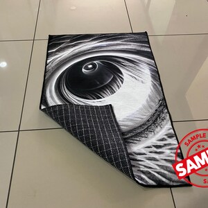 May include: Black and white rug with a large eye design. The rug has a black and white checkered backing.