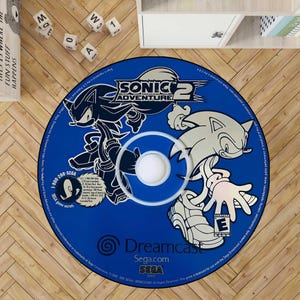May include: A blue circular rug featuring the Sonic Adventure 2 video game logo and characters. The rug has a white and black design of Sonic the Hedgehog. The rug is on a wooden floor.