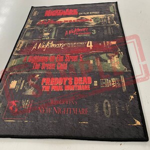 May include: A black and red rug with a design of VHS tapes featuring the Nightmare on Elm Street movie franchise. The rug features the titles "A Nightmare on Elm Street", "A Nightmare on Elm Street 3", "A Nightmare on Elm Street 4", "A Nightmare on Elm Street 5: The Dream Child", "Freddy's Dead: The Final Nightmare", and "Wes Craven's New Nightmare".