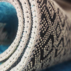 May include: Close-up of a rolled-up rug with a white, textured border. The rug features a blue and white pattern with a black and white geometric design. The rug is made of a soft, plush material.