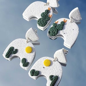 May include: A pair of white ceramic earrings with a desert theme. Each earring features a white arch with a yellow sun and green cactus plants.