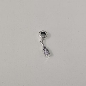 Puede incluir: Un charm de plata con un diseño de botella de champán. El charm presenta una parte superior pequeña y redonda con el texto "925 ALE" y una forma de botella colgante cubierta de pequeñas piedras brillantes. El charm está sobre un fondo blanco.