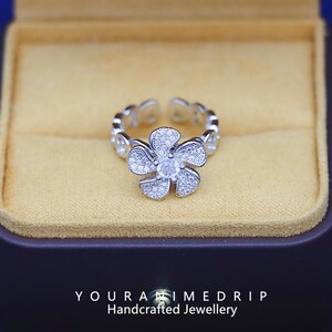 May include: A silver ring with a flower design set with clear cubic zirconia stones. The ring is in a small, velvet-lined box. The box has the text "YOUR ANIMEDRIP Handcrafted Jewellery" on the bottom.