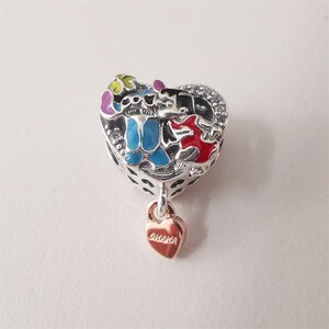 May include: A silver charm with a heart-shaped design featuring colorful cartoon characters. The charm has a small, rose-gold heart pendant with the word "Ohana" engraved on it. The charm is likely for a bracelet or necklace.
