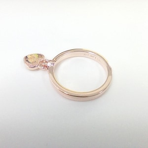 May include: Rose gold ring with a heart-shaped charm. The charm has a yellow center and a decorative border. The ring is set against a white background, showcasing its delicate design and color.