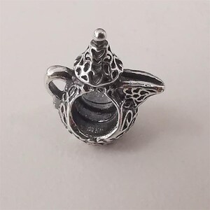 Puede incluir: Un charm plateado detallado con forma de tetera. El charm presenta intrincados patrones calados en el cuerpo, la tapa y el pico. Un pequeño botón redondo se encuentra en la tapa. El charm tiene un lazo para sujetarlo a una pulsera o collar.