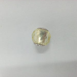 Puede incluir: Una cuenta facetada de color amarillo claro con bandas plateadas en cada extremo. La cuenta tiene un diseño geométrico y parece estar hecha de vidrio o un material similar. Las bandas tienen texto grabado. La cuenta es un componente de joyería.