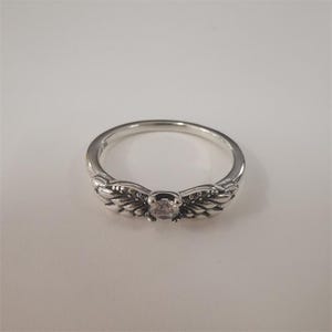 May include: A silver ring with a central clear gemstone and wing-shaped accents. The ring has a delicate design, with detailed feathering on the wings. The ring is set against a white background.