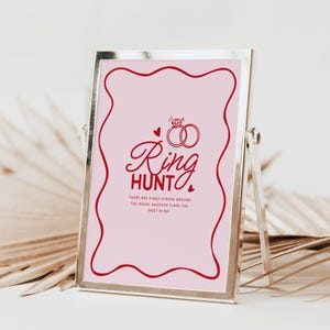 K&ouml;nnte beinhalten: Ein rosa Schild mit rotem Rand und dem Text "Ring Hunt" in rot. Das Schild hat auch ein rotes Herz und zwei rote Ringe mit einem Diamanten. Der Text unter den Ringen lautet "Es sind Ringe im Raum versteckt. Wer die meisten findet, gewinnt!"