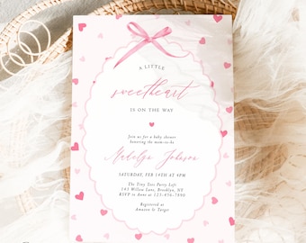 May include: A baby shower invitation in pink and white. The invitation has the text "A Little Sweetheart Is On The Way" and event details. Pink hearts and a pink bow decorate the invitation. The invitation is for a baby shower honoring the mom-to-be.