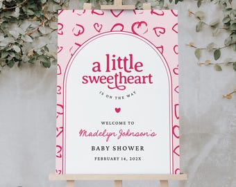 May include: A pink baby shower welcome sign on an easel. The sign features the text "a little sweetheart is on the way" in red script. The sign also includes the name "Madelyn Johnson's" and the date "February 14, 202X".