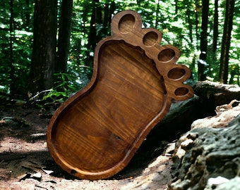 May include: A wooden serving tray shaped like a human foot. The tray is made of dark brown wood and has a smooth, polished finish. The toes are individually carved, and the tray is set against a natural forest backdrop.