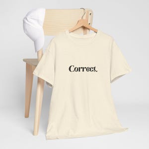 May include: A cream-coloured short-sleeve t-shirt with a speckled texture and the word Correct. printed in black serif font on the chest, accompanied by a plain white baseball cap hanging on the back of a light-coloured wooden chair.