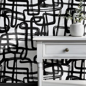 May include: A white wall covered in wallpaper featuring a black hand-painted brushstroke pattern of intersecting lines and loops. A white wooden console table with a single drawer and a small white vase with green foliage sits in front.