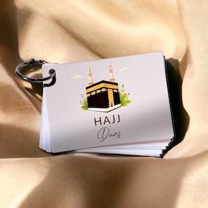 May include: A small flipbook with a silver metal ring. The cover features an illustration of the Kaaba with the words "HAJJ Duas" in a script font. The background is a light beige.