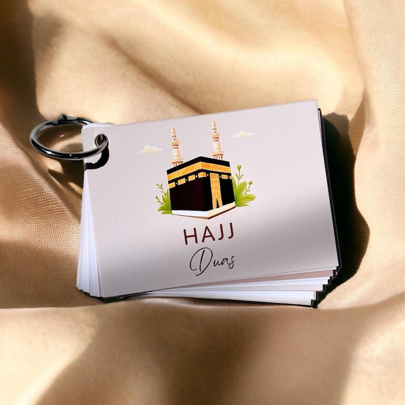 May include: A small flipbook with a silver metal ring. The cover features an illustration of the Kaaba with the words "HAJJ Duas" in a script font. The background is a light beige.