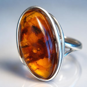May include: A silver ring featuring a large, oval-shaped, translucent amber gemstone. The amber displays deep orange and brown hues with visible internal inclusions. The stone is set within a polished silver bezel and a split-shank band design.