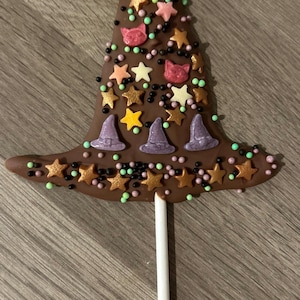 May include: A milk chocolate lollipop shaped like a pointed hat, decorated with edible star-shaped sprinkles in gold, yellow, and pink, small purple hat-shaped candies, pink cat-face shapes, and tiny black and green round sugar pearls.