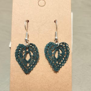 May include: A pair of handmade, dark teal crochet heart-shaped earrings. The earrings are suspended from silver-colored fish hook ear wires. The intricate openwork design of the hearts is visible against the neutral background.
