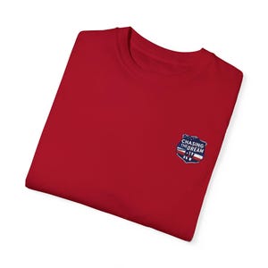 Puede incluir: Camiseta roja con un parche azul y blanco que dice "Chasing the Dream - IV - RW"