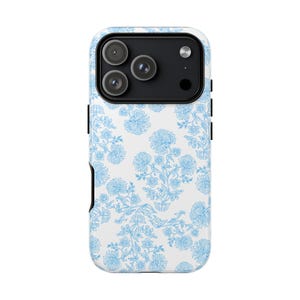 Op de afbeelding: Een smartphonehoesje met een witte achtergrond en een herhalend lichtblauw bloemenpatroon. Het hoesje heeft een zwart cameramodule aan de bovenkant met drie ronde lenzen en een flitser. Het ontwerp bedekt de achterkant van het toestel.