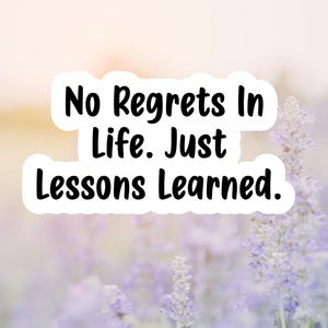 May include: A sticker with the text "No Regrets In Life. Just Lessons Learned." in a black font, set against a blurred background of lavender flowers and a soft, warm light.