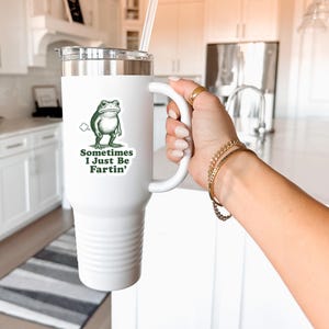 May include: White insulated tumbler with a handle, a silver lid, and a clear straw. The tumbler features a green cartoon frog graphic with the text "Sometimes I Just Be Fartin'." The tumbler is held by a person.