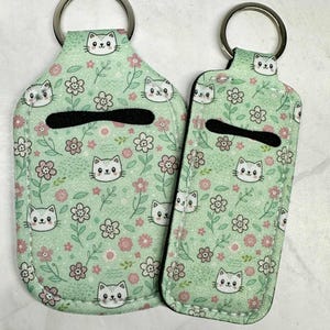 May include: Two keychains with a mint green floral and cat pattern. The keychains have a black opening and a silver key ring. The pattern includes pink flowers, white cat faces, and green leaves.