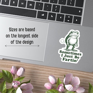 May include: A green frog sticker on a laptop with the text "Sometimes I Just Be Fartin'". The laptop also displays the text "Sizes are based on the longest side of the design". The laptop is on a wooden surface with pink flowers.