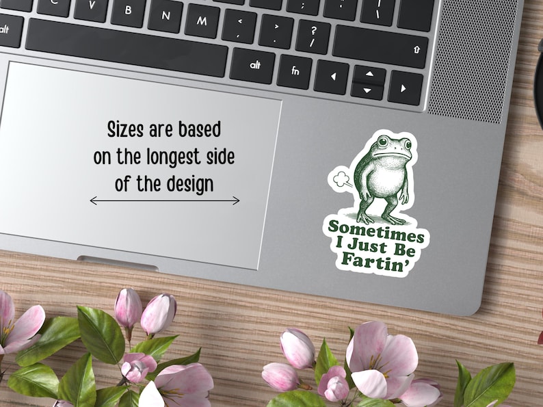 May include: A green frog sticker on a laptop with the text "Sometimes I Just Be Fartin'". The laptop also displays the text "Sizes are based on the longest side of the design". The laptop is on a wooden surface with pink flowers.