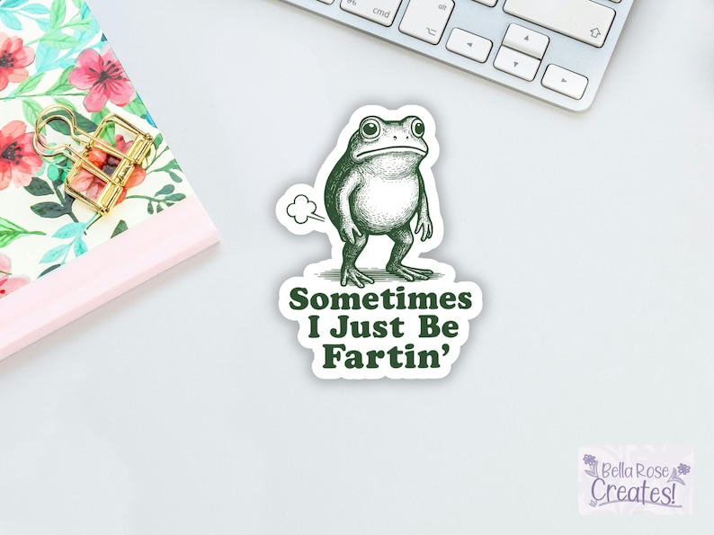 May include: A green and white sticker featuring a cartoon frog with the text "Sometimes I Just Be Fartin'". The sticker is on a white surface near a keyboard and a floral notebook.