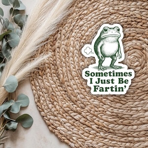 May include: A whimsical sticker featuring a green frog with a comical expression, emitting a puff of gas. The sticker includes the text "Sometimes I Just Be Fartin'" in a playful font. The sticker is set against a woven background.