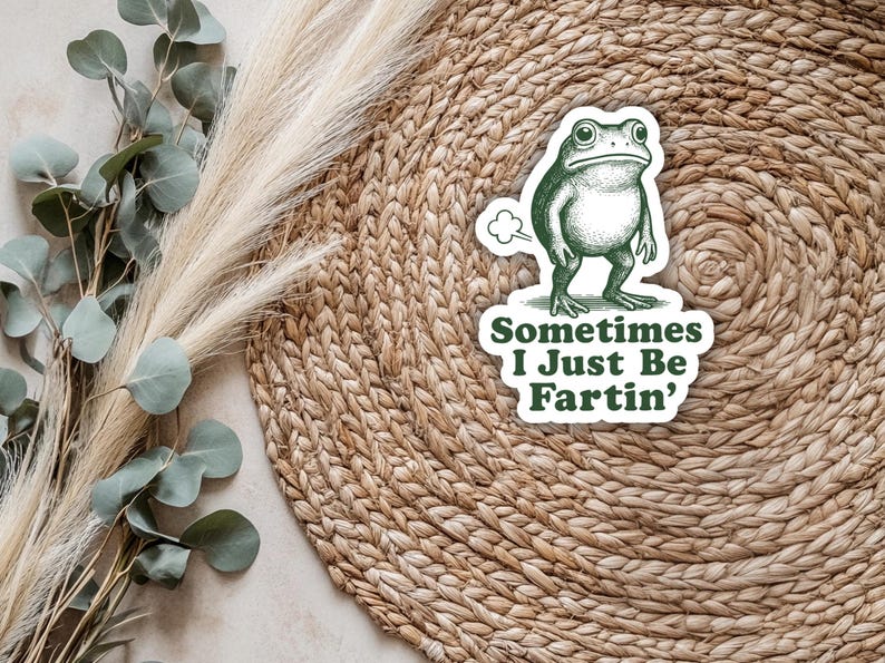 May include: A whimsical sticker featuring a green frog with a comical expression, emitting a puff of gas. The sticker includes the text "Sometimes I Just Be Fartin'" in a playful font. The sticker is set against a woven background.