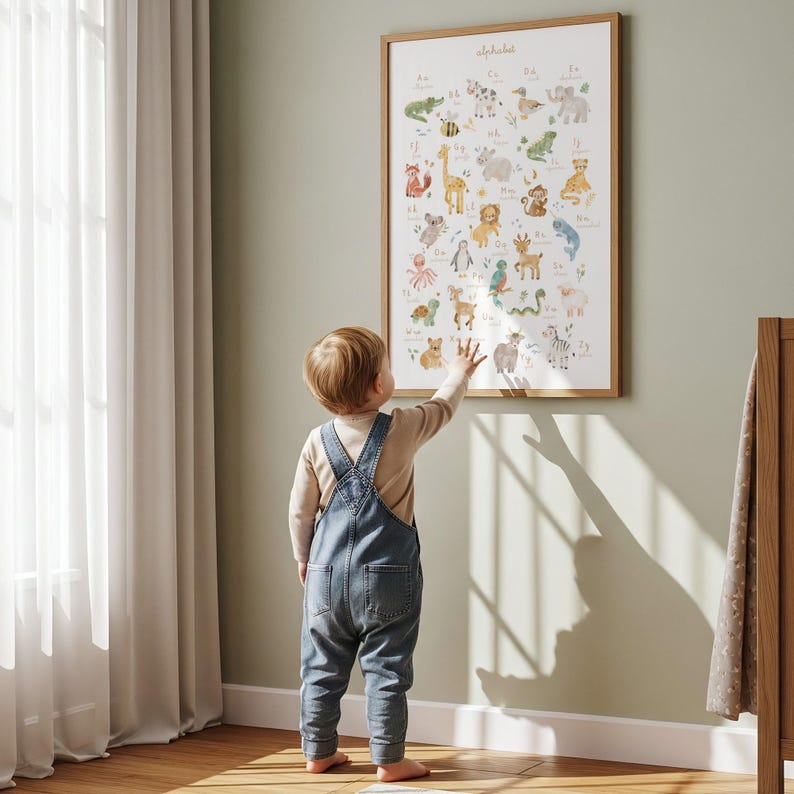 Little boy in denim overalls standing with his back turned, pointing at a framed ABC watercolor animals poster on a sage green nursery wall. Bright sunlight casting soft window shadows in a modern minimalist kids bedroom.