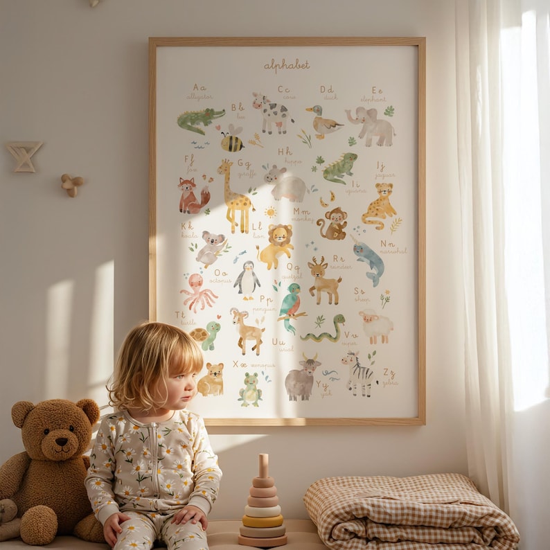 Sweet toddler girl in pajamas sitting on the floor next to a teddy bear and wooden toy pyramid, looking at a large framed watercolor animal alphabet chart hanging on a sunlit nursery wall. Warm, aesthetic scandinavian style playroom.