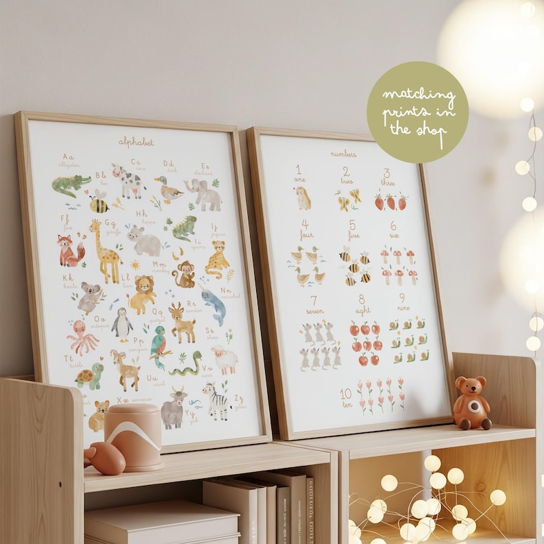 Matching prints set for kids room. Two framed posters sitting on a wooden shelf with fairy lights: the animal alphabet chart and a matching numbers poster. Green sticker reads "matching prints in the shop" for gallery wall inspiration.
