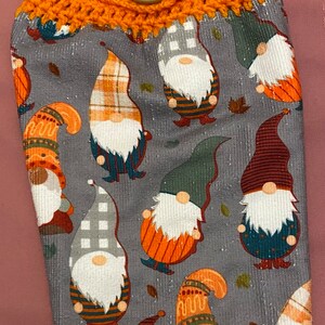 May include: Orange crocheted dish towel holder with a wooden button. The towel is gray with a pattern of gnomes wearing different colored hats and outfits.