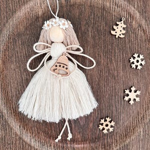 May include: A handmade Christmas ornament in the shape of an angel, featuring a wooden head, beige yarn hair and dress, and a wooden bell. The angel is adorned with a white flower crown and is accompanied by wooden snowflakes and a Christmas tree.
