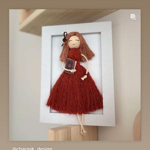 Puede incluir: Muñeca hecha a mano en un marco blanco. La muñeca tiene un vestido rojo, cabello castaño y un libro pequeño. El cabello de la muñeca es castaño y tiene un lazo negro. La muñeca sostiene un libro con una cubierta marrón.