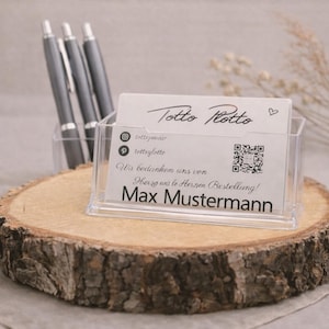 Könnte beinhalten: Ein klarer Acryl-Visitenkartenhalter mit dem Text "Totto Rotto" und "Max Mustermann" auf einer weißen Karte. Der Halter steht auf einer rustikalen Holzscheibe, mit drei grauen Stiften in einem transparenten Behälter dahinter.