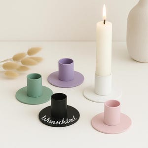 Op de afbeelding: Een verzameling kleurrijke kaarshouders in pasteltinten groen, paars, roze en zwart. Eén houder heeft het woord "Wunschtext" in witte letters. Een witte kaars brandt in een witte houder. Decoratieve woondecoratie.