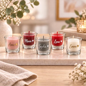 Peut inclure: Cinq petits bougeoirs en verre contenant chacun une bougie en cire colorée. Les bougies sont rose, rouge foncé, grise, rouge vif et blanche. Chaque verre porte un prénom écrit en lettres cursives : Anna, Marie, Sophie, Lena et Emma, avec un cœur.