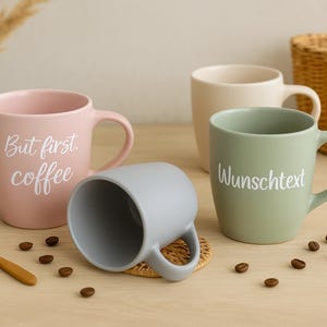 Könnte beinhalten: Ein Set aus vier Keramiktassen in Pastellrosa, Salbeigrün, Hellgrau und Creme. Die rosa Tasse trägt den weißen Schriftzug "But first, coffee", während auf der salbeigrünen Tasse der weiße Text "Wunschtext" steht. Ein kleiner Holzlöffel liegt daneben.